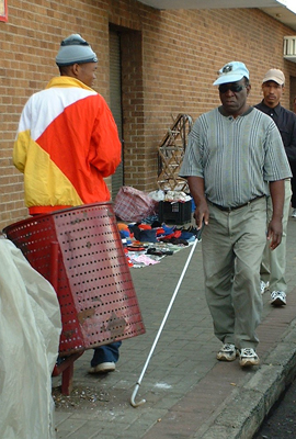 The same man from the last photo is walking along a sidewalk with his hand centered in front of his waist, and reaching on the ground ahead of him with his long white cane with bundu basher tip.